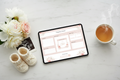 Digital pregnancy journal planner on a tablet, flat lay on white marble with baby shoes, soft pink flowers and herbal tea