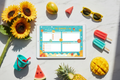 Digital summer planner for kids on a tablet, flat lay on white marble with sunflowers, popsicles, beach bucket, sunglasses and tropical fruit